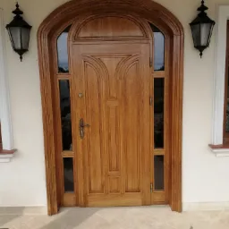 Wejściowe drzwi z drewna, o łukowatym kształcie, z przeszkleniami po bokach, zamontowane w jasnej elewacji, oświetlone przez dwie czarne lampy zewnętrzne.