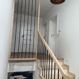 Wnętrze domu w trakcie wykańczania z widocznymi schodami z jasnego drewna z czarnymi balustradami, niedokończoną ścianą i sufitem oraz nowoczesną lampą wiszącą.