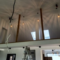 FIRMA MONTERSKO-USŁUGOWA IRO-TECH - Wnętrze domu w trakcie wykańczania: drewniane słupy podtrzymujące strop, schody, drabina aluminiowa, wiszące lampy z żarówkami Edisona, instalacja elektryczna i okna dachowe.