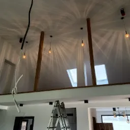 Wnętrze domu w trakcie wykańczania: drewniane słupy podtrzymujące strop, schody, drabina aluminiowa, wiszące lampy z żarówkami Edisona, instalacja elektryczna i okna dachowe.