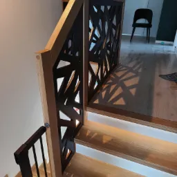 Nowoczesne schody z dębowymi stopniami podświetlonymi od spodu, balustrada z drewnianą ramą i czarnym, ażurowym wypełnieniem geometrycznym, rzucającym dekoracyjne cienie na podłogę.