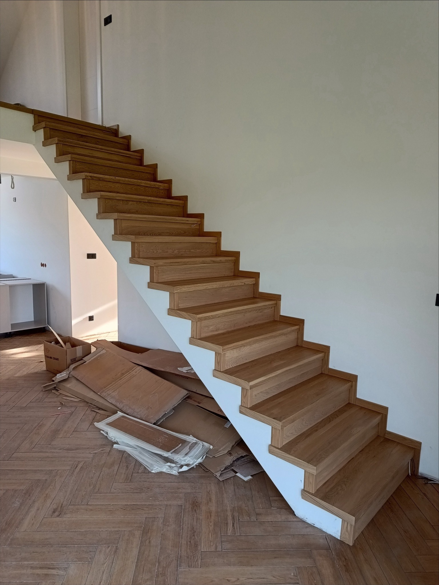 Proste, drewniane schody z białymi policzkami, widok z dołu, na podłodze kartony po montażu, parkiet w jodełkę.