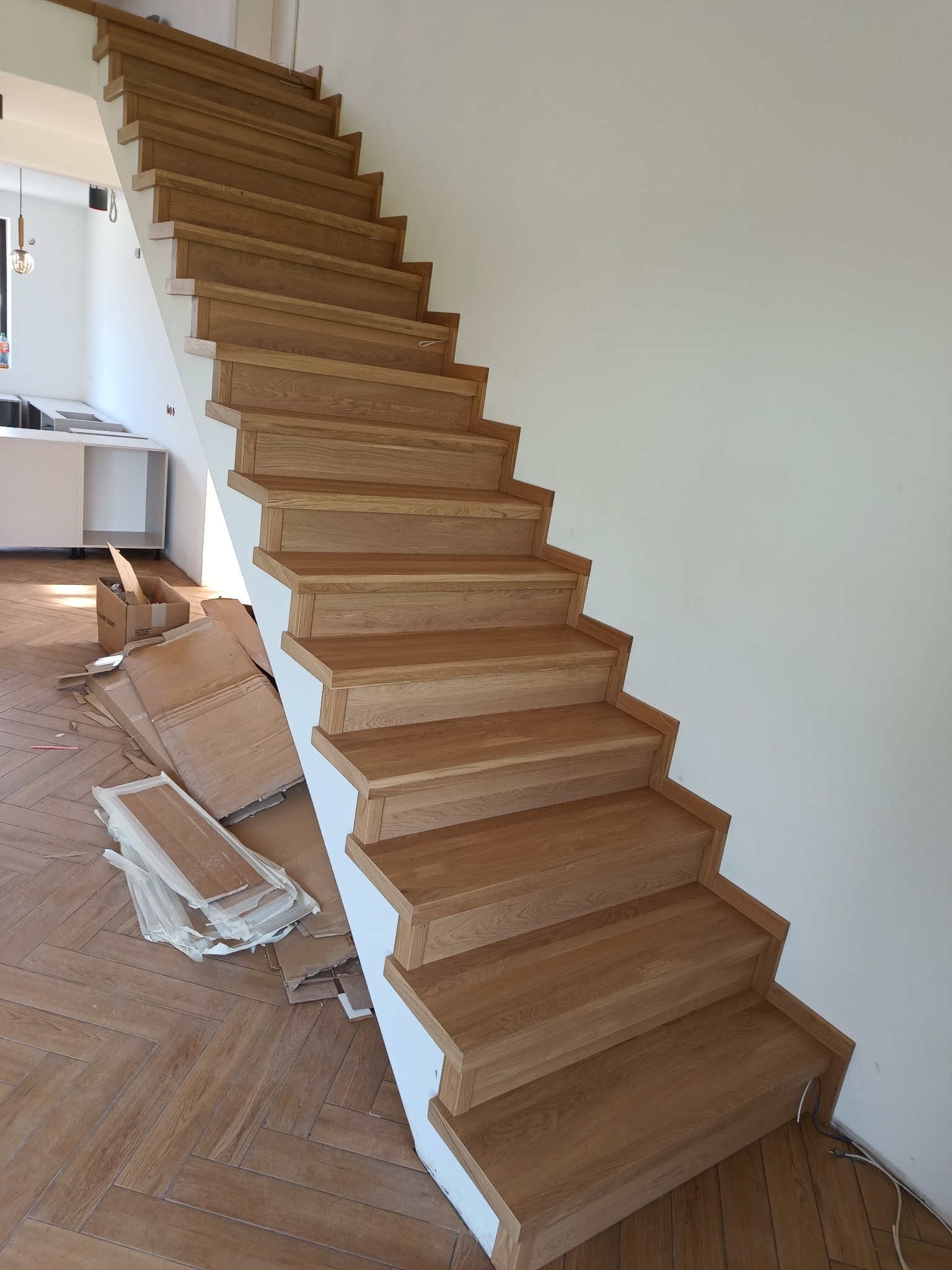 Nowe dębowe schody z białymi podstopnicami, widoczne kartony po montażu i parkiet ułożony w jodełkę.