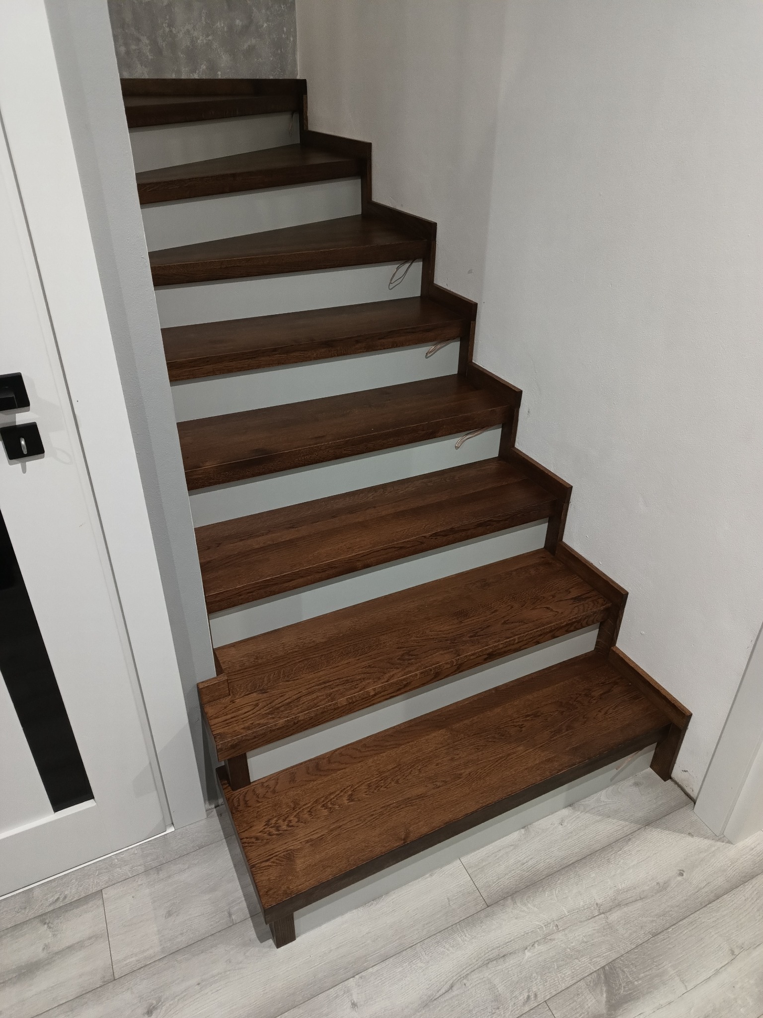 Wewnętrzne schody z ciemnego drewna, osadzone na szarych podstopnicach, widoczne od dołu, przy białej ścianie i obok białych drzwi z czarnym akcentem.