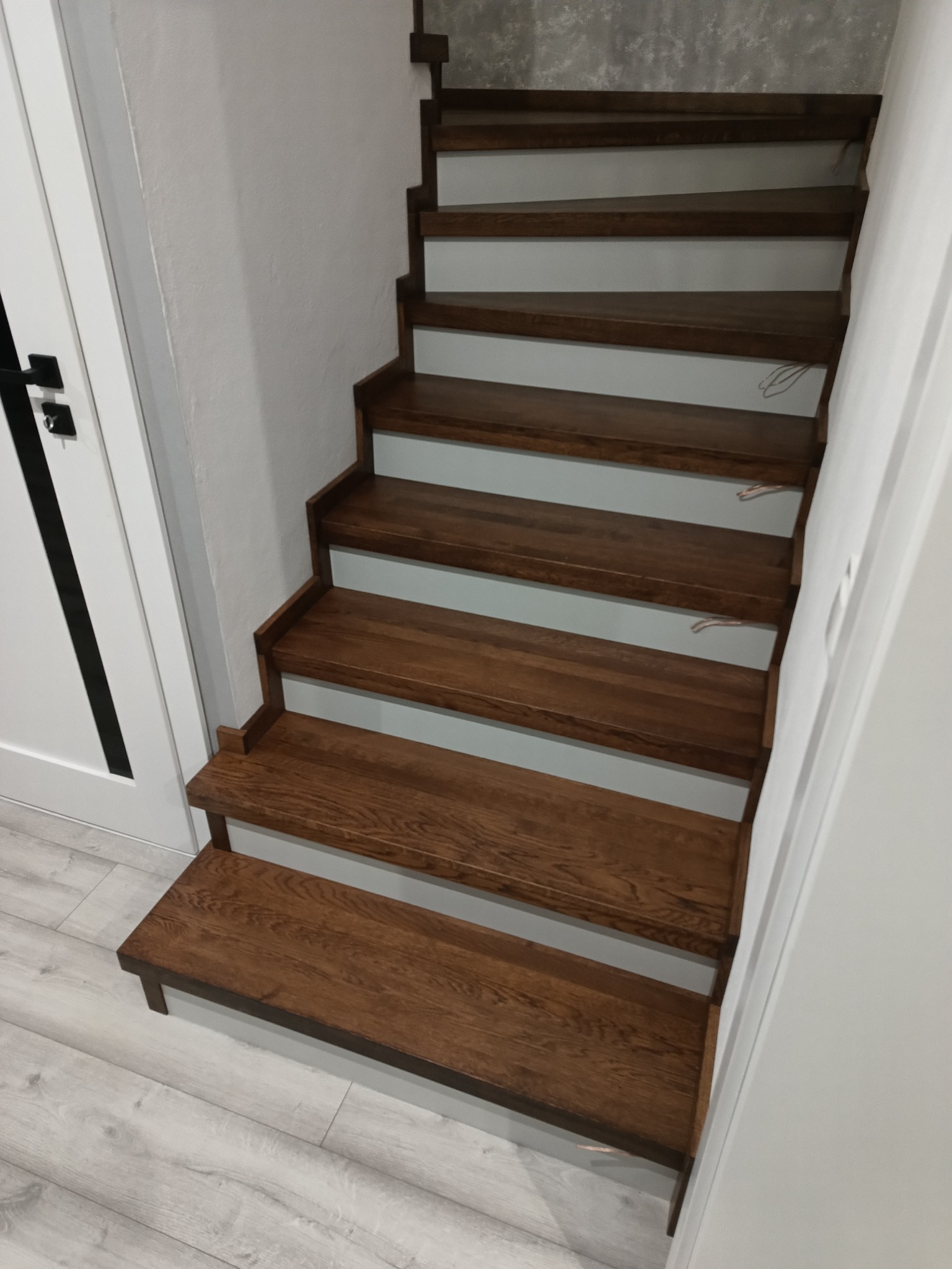 Wewnętrzne schody z ciemnego drewna z białymi podstopnicami, widok z góry, przy ścianie i białych drzwiach z czarną klamką.