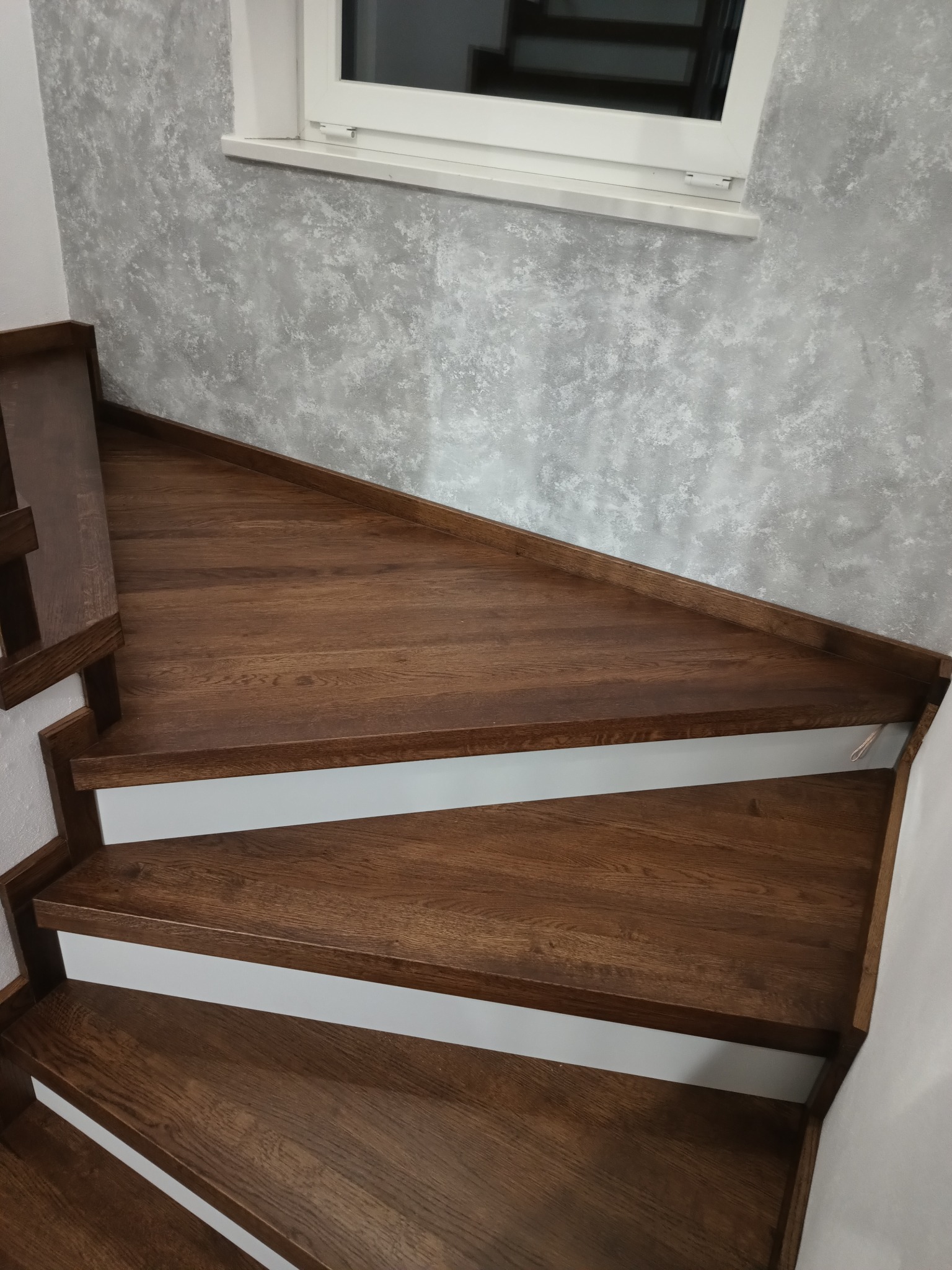 Drewniane schody w odcieniach brązu, z białymi podstopnicami, prowadzące w górę, widoczne z góry, przy szarej ścianie z efektem dekoracyjnym i oknem w tle.