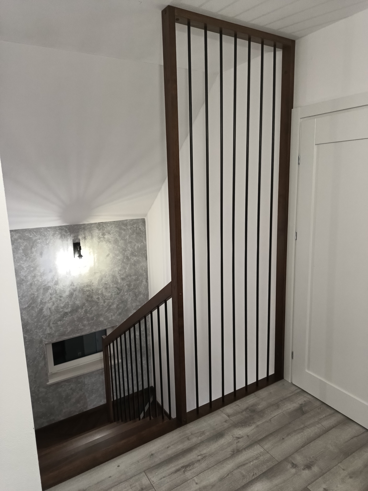 Nowoczesna klatka schodowa z drewnianymi schodami i balustradą z czarnych, pionowych prętów, oddzielona od reszty pomieszczenia drewnianą ścianką działową, widok z góry na dół.