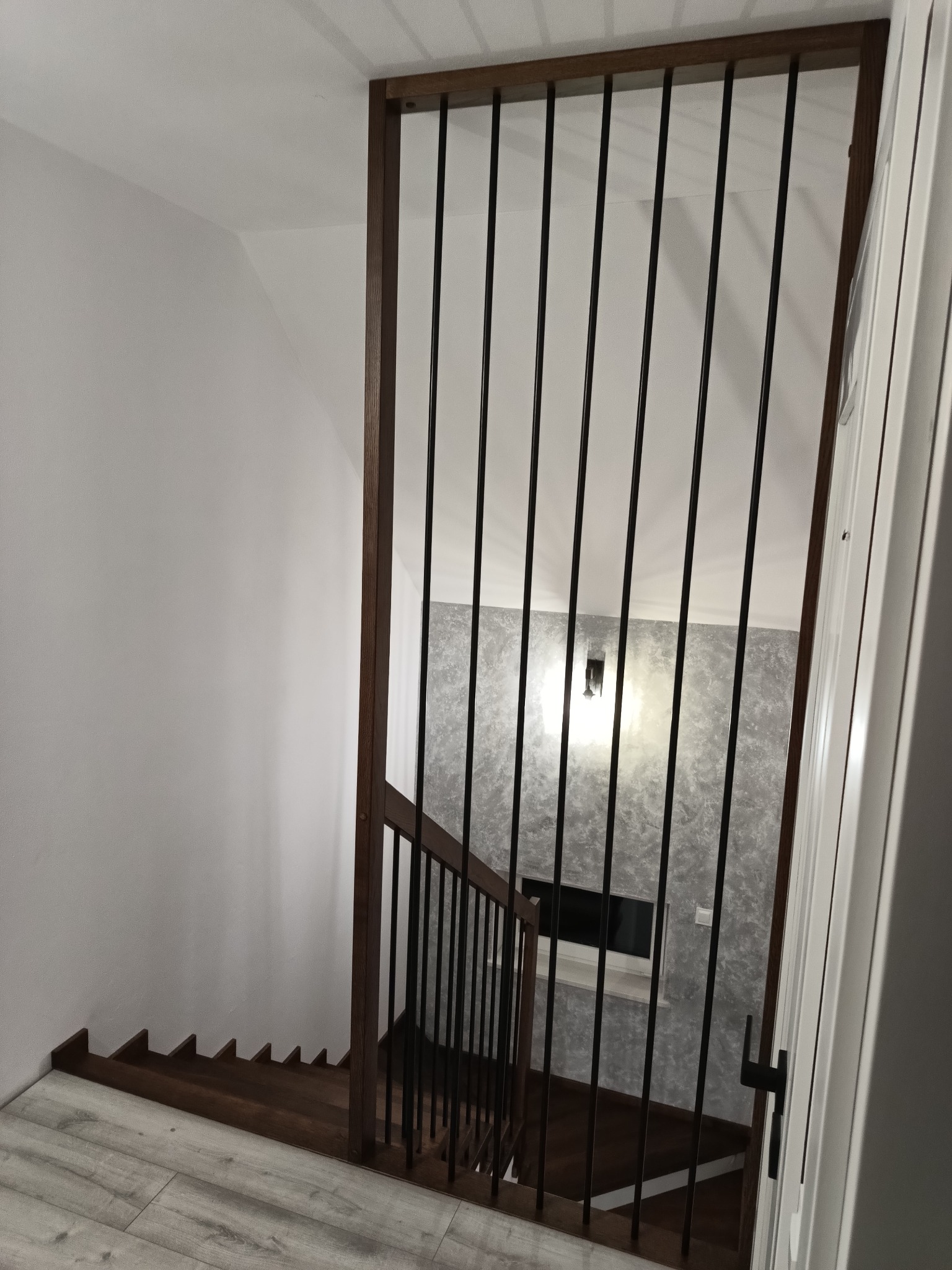 Widok z góry na drewniane schody z czarnymi, metalowymi balustradami, z drewnianą ramą, prowadzące w dół do pomieszczenia z szarą ścianą i oświetleniem punktowym.