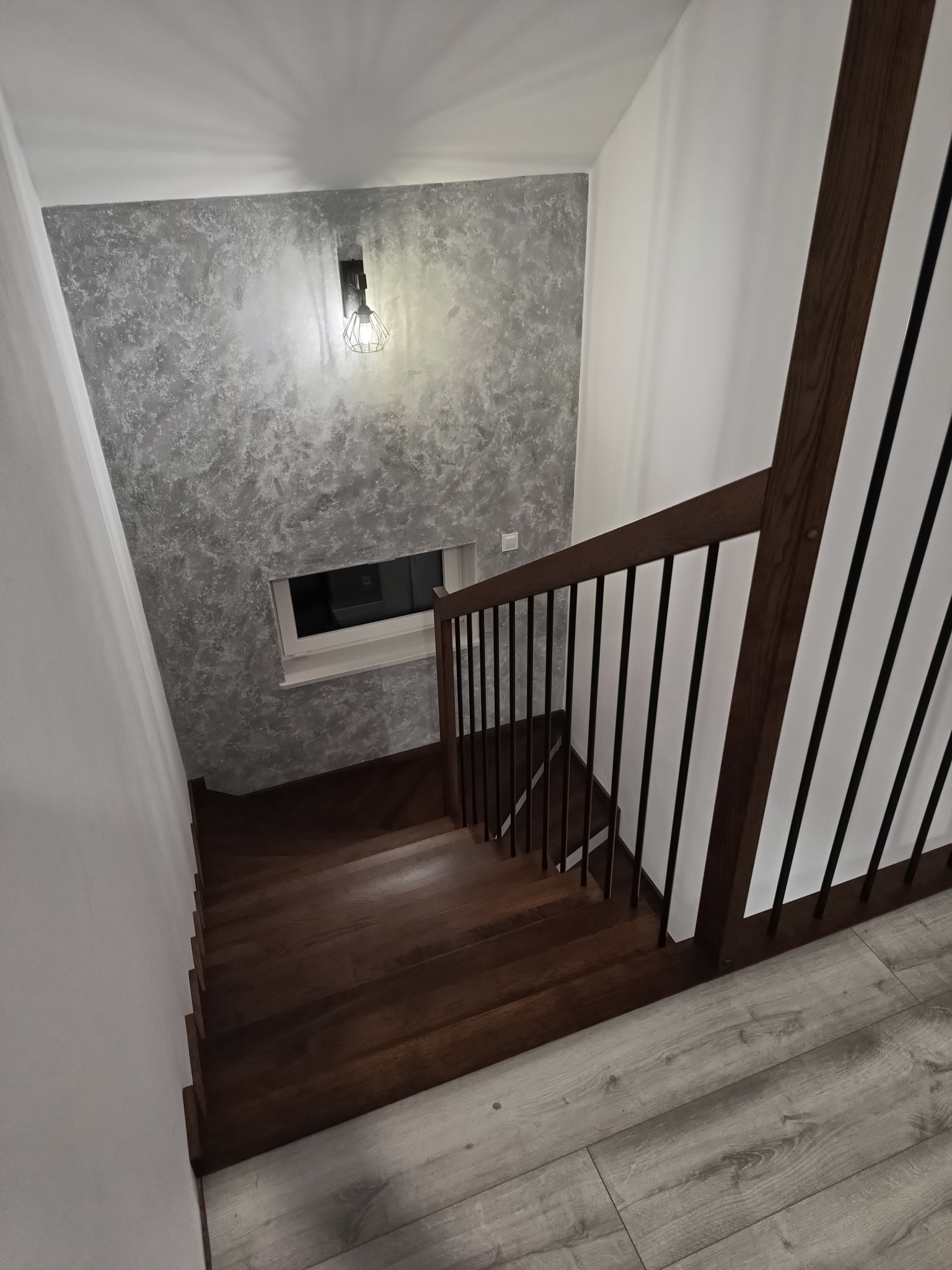 Wnętrze z drewnianymi schodami w ciemnym odcieniu, czarne metalowe balustrady, ściana z efektem dekoracyjnym oraz widok na okno i oświetlenie w industrialnym stylu.