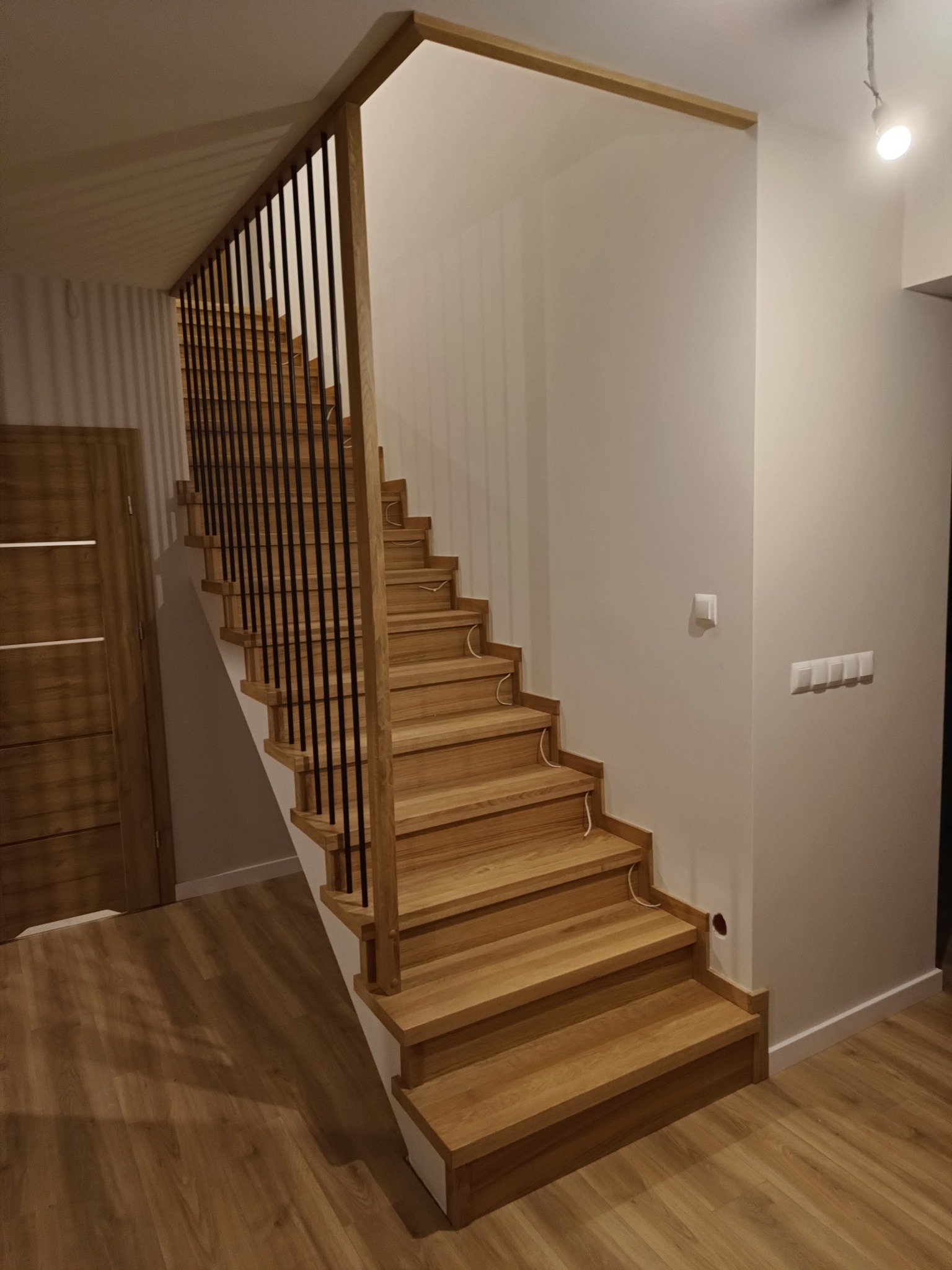 Nowoczesne schody z drewnianymi stopniami i czarną, metalową balustradą, oświetlone ciepłym światłem, widoczne kable elektryczne przy stopnicach, drewniana podłoga w odcieniach brązu.