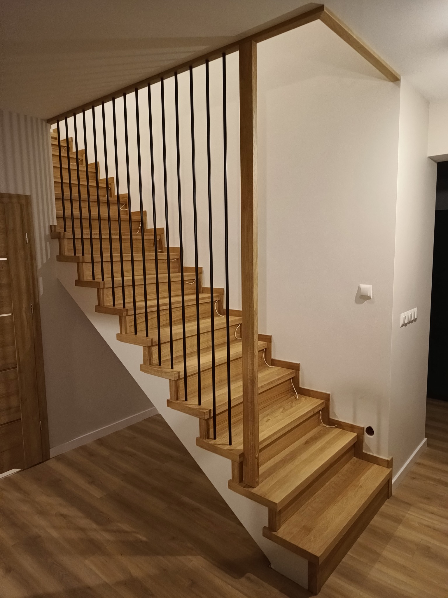 Jasne, drewniane schody z czarnymi, metalowymi balustradami w nowoczesnym wnętrzu, widoczne przewody elektryczne przy schodach.