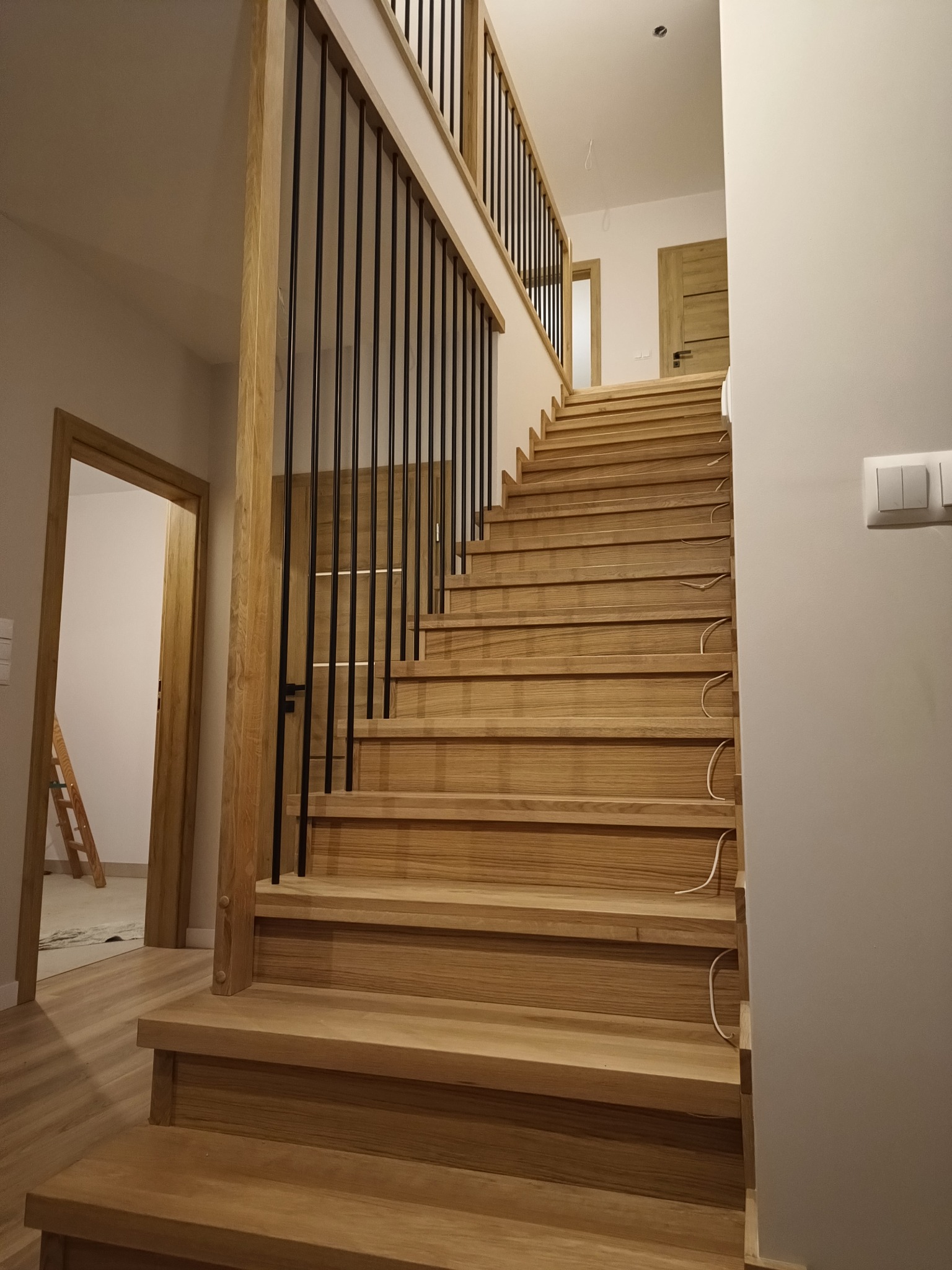 Jasne, drewniane schody z czarną, minimalistyczną balustradą, prowadzące do góry w nowoczesnym wnętrzu. Widoczne kable wystające spod stopni. W tle drzwi i drabina.