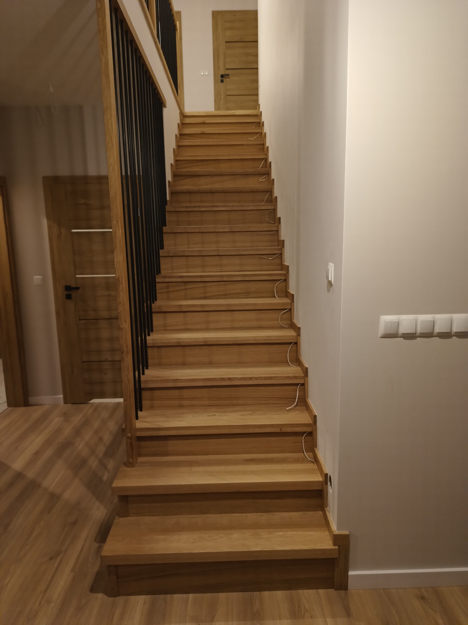 Proste, drewniane schody z czarną balustradą w nowoczesnym wnętrzu, widoczne wystające kable instalacji elektrycznej.