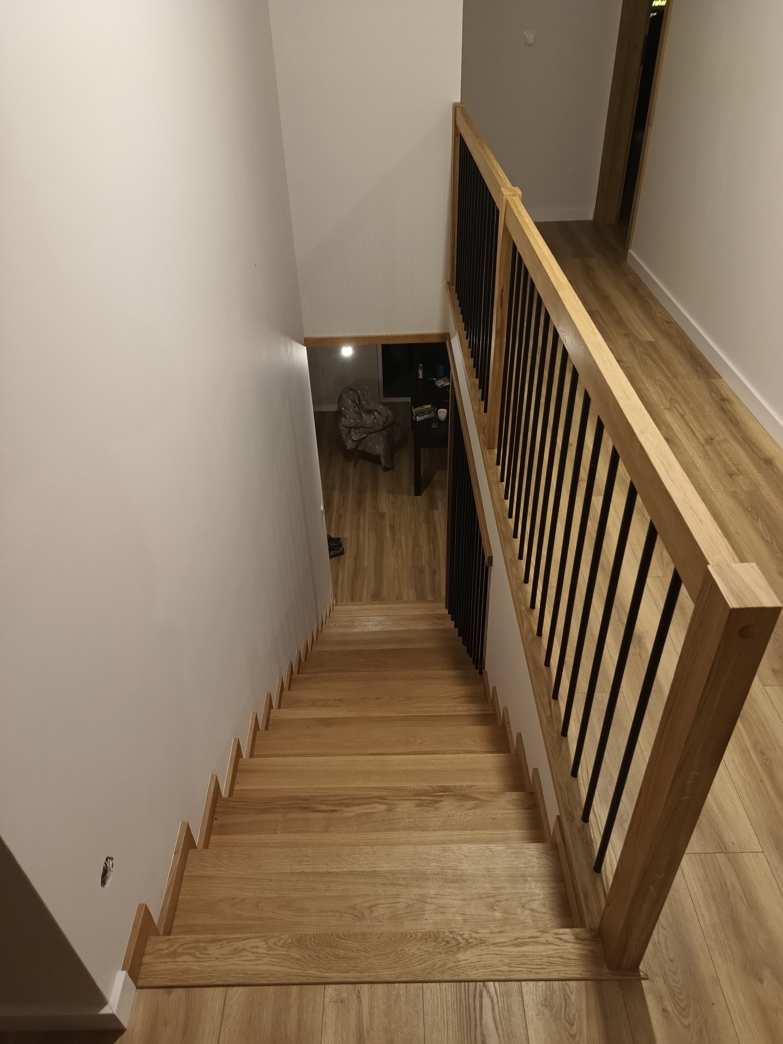 Drewniane schody z balustradą z czarnymi metalowymi prętami, widok z góry, jasne drewno, minimalistyczny design, widoczne oświetlenie w tle.