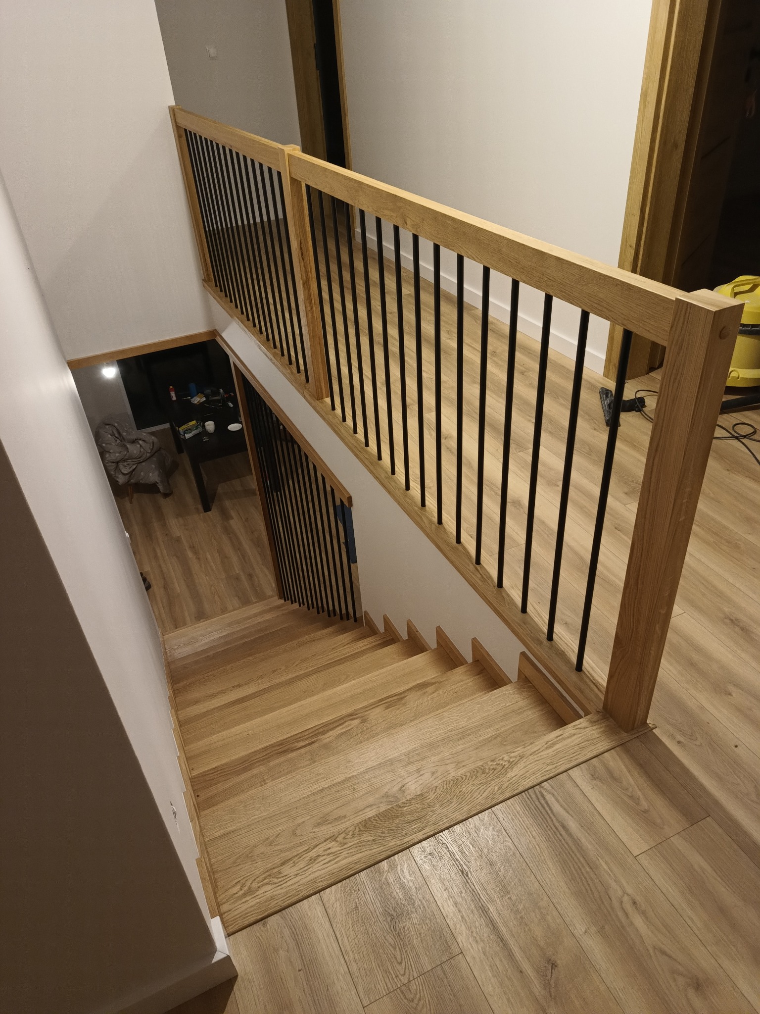 Jasne, drewniane schody z czarnymi, metalowymi balustradami, widok z góry na dół, z widocznym fragmentem salonu w tle.