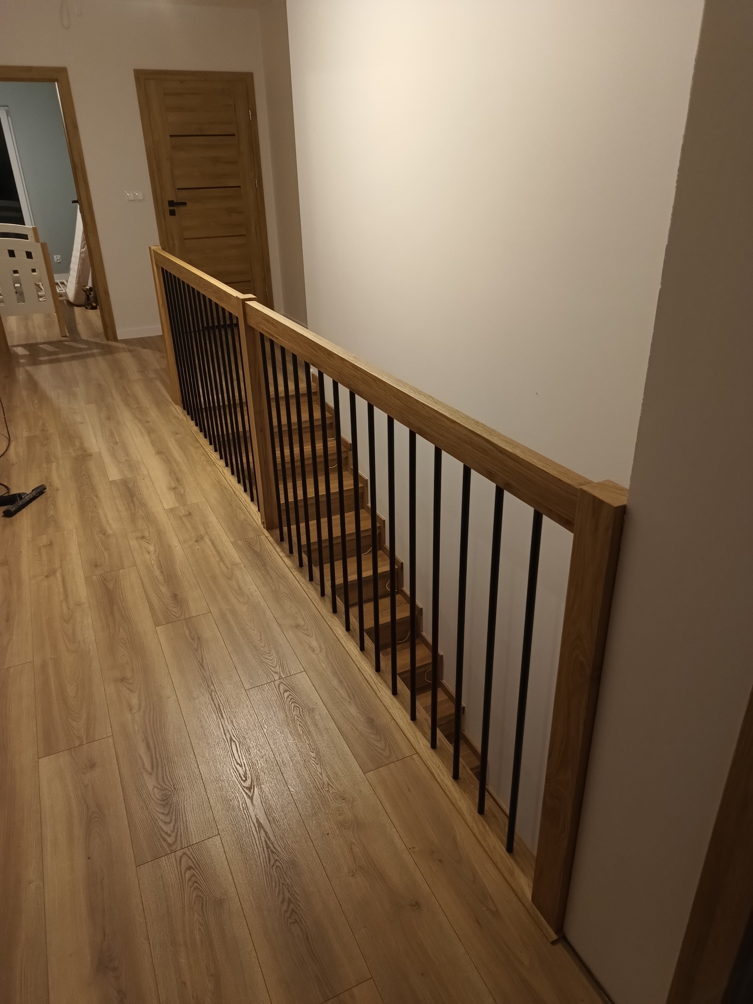 Nowoczesne schody z drewnianymi stopniami i balustradą z czarnymi, metalowymi prętami, widok z góry korytarza z drewnianą podłogą i drzwiami.