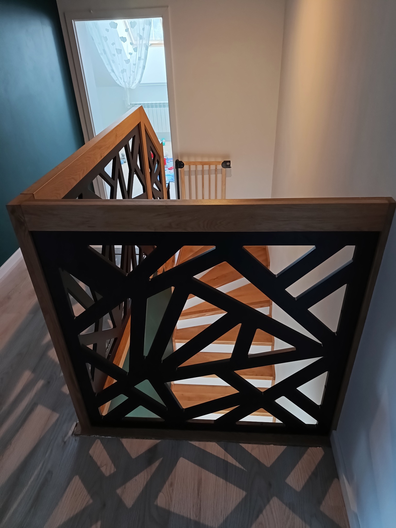 Nowoczesna balustrada schodowa z drewnianą ramą i geometrycznym wypełnieniem z czarnych, przecinających się elementów, widziana z góry, z widocznymi fragmentami drewnianych schodów w tle.