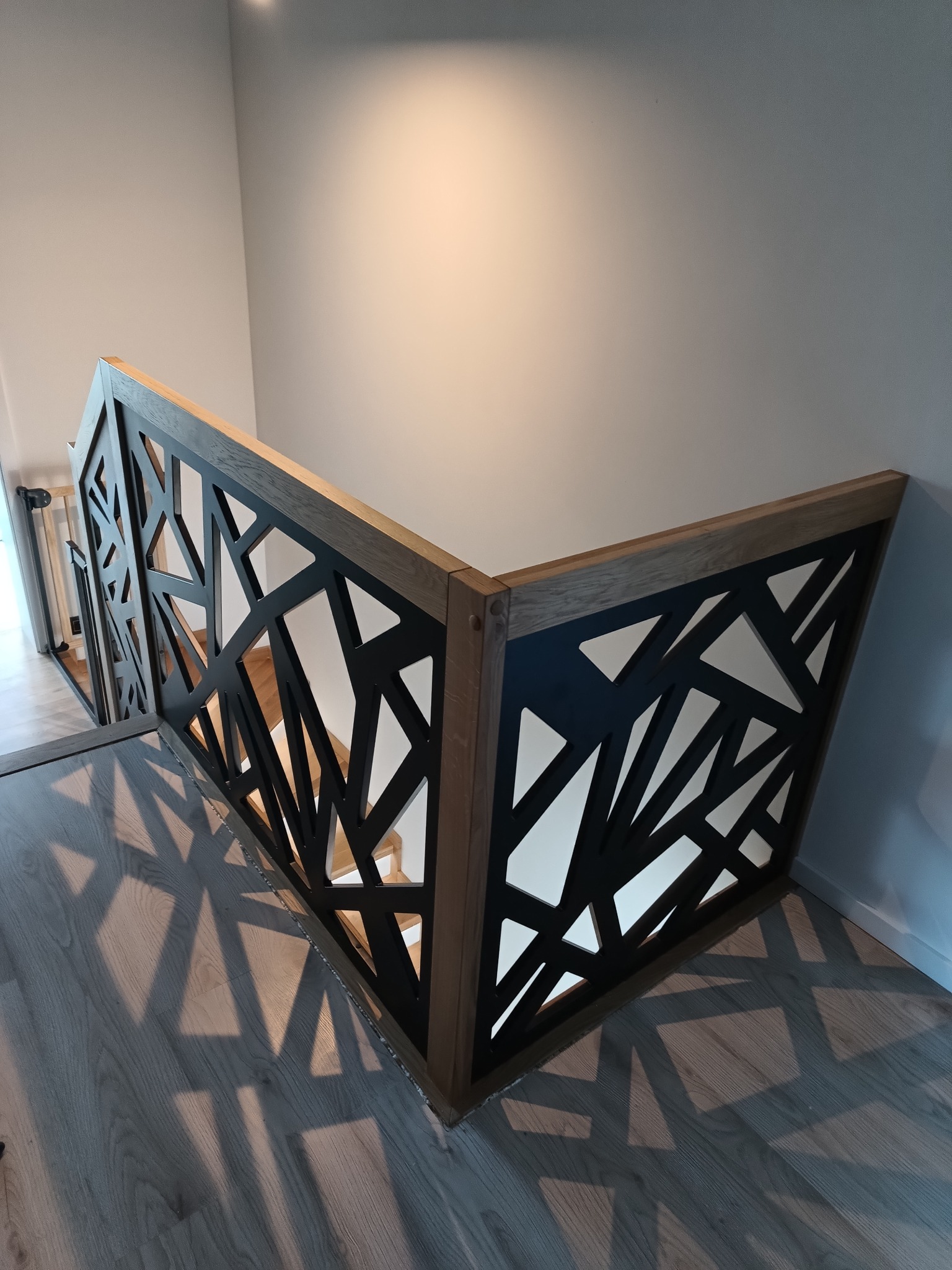Nowoczesna balustrada schodowa z drewnianą ramą i czarnym, geometrycznym wypełnieniem, rzucająca cienie na podłogę z paneli w jasnym wnętrzu.