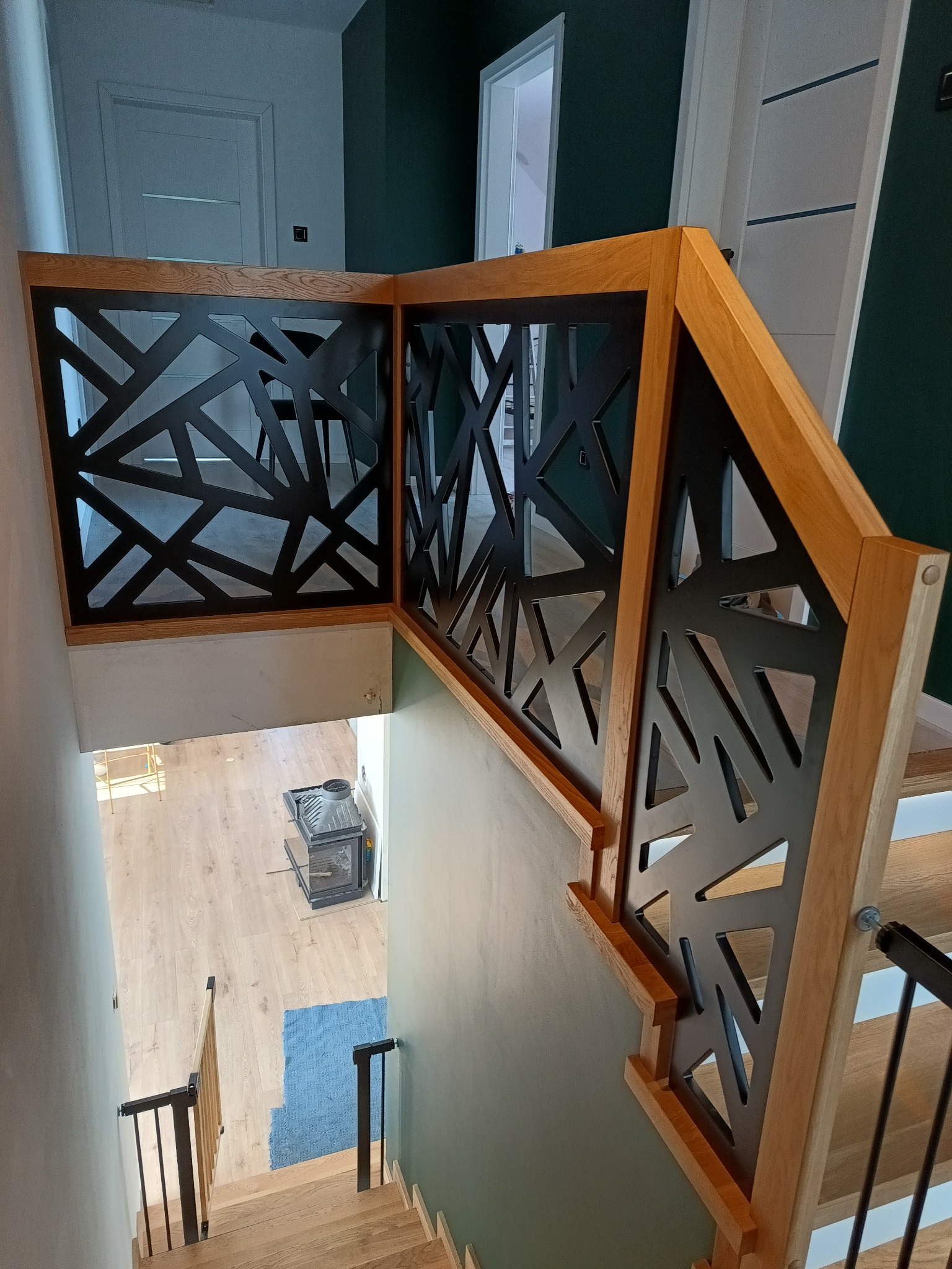 Nowoczesna balustrada z drewna i czarnego metalu o geometrycznym wzorze, widok z góry na schody z oświetleniem LED, w tle jasna podłoga i piec.