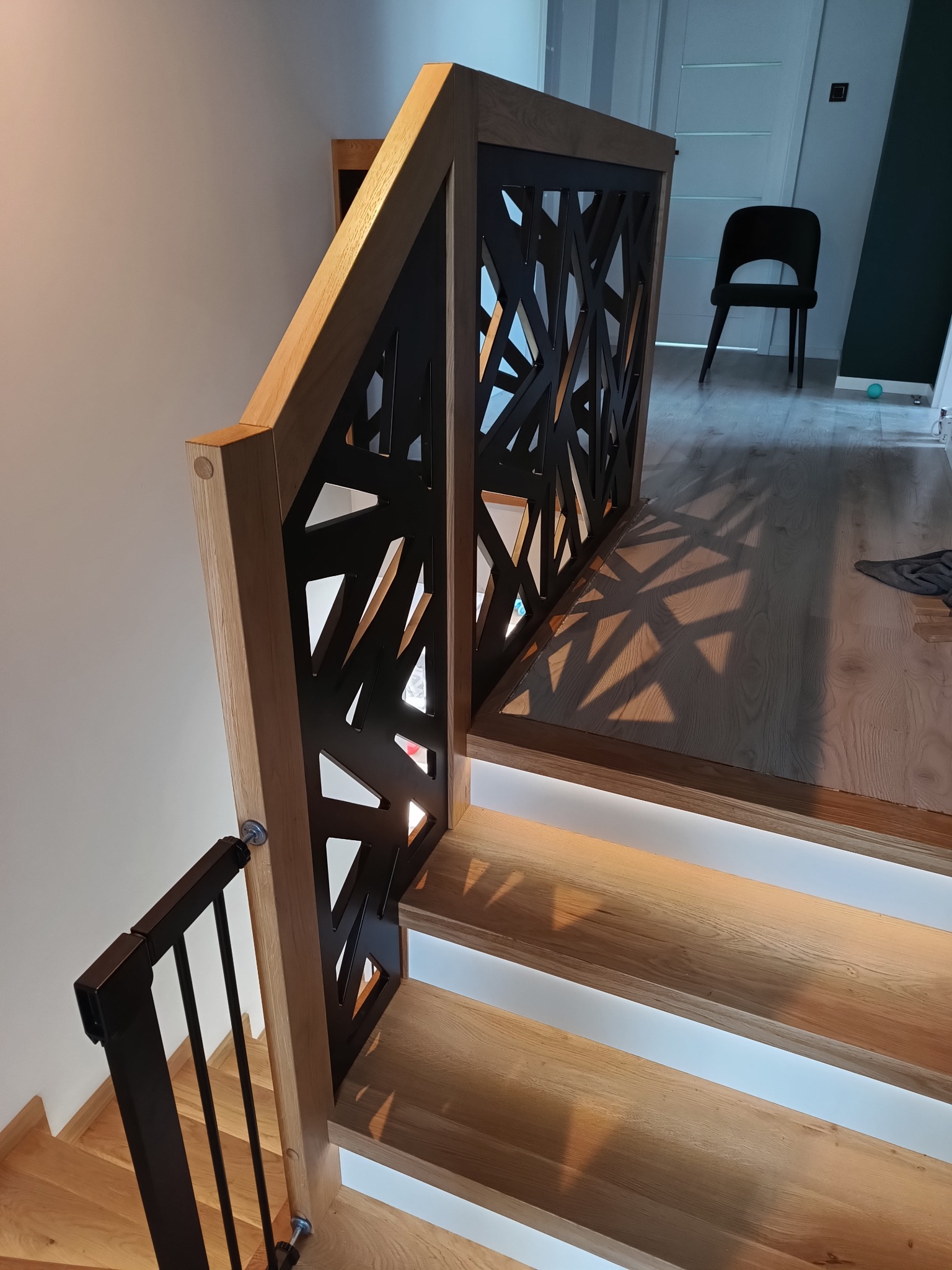 Nowoczesne schody z dębowymi stopniami podświetlonymi od spodu, balustrada z drewnianą ramą i czarnym, ażurowym wypełnieniem geometrycznym, rzucającym dekoracyjne cienie na podłogę.