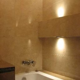 WM-Bud - Wykończona łazienka z beżowymi płytkami imitującymi kamień, biała wanna z chromowaną baterią, oświetlenie punktowe w półce ściennej.