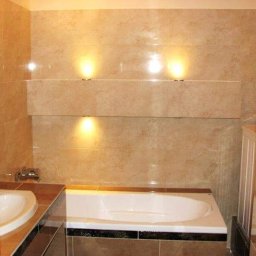 WM-Bud - Wykończona łazienka z beżowymi płytkami imitującymi marmur, podświetlanymi niszami, białą wanną z czarnym obramowaniem i umywalką z brązowym blatem.