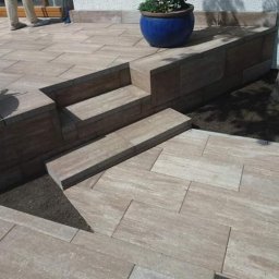 WM-Bud - Wykończony taras z kostki brukowej o nieregularnym wzorze, z niskim murkiem i schodkami, donica z kwiatami na murku, widoczne fragmenty drzwi i drzewa.