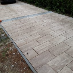 WM-Bud - Ułożona kostka brukowa w odcieniach beżu i szarości, tworząca podjazd z widocznym odpływem liniowym. Z boku widoczne krzewy i trawnik z elementami ściółki leśnej.