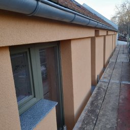 WM-Bud - Fragment elewacji budynku z oknem w zielonej ramie i szarym parapetem, widoczny z rusztowania, z fakturą baranka w odcieniach beżu i pomarańczy.