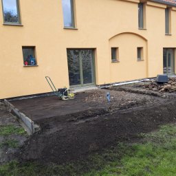 WM-Bud - Przygotowanie terenu pod taras przy żółtej elewacji domu, widoczna ubijarka, wykop, obrzeża betonowe i sterta gruzu.