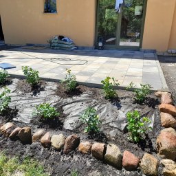 WM-Bud - Układanie kostki brukowej na tarasie przy domu z przygotowanym miejscem na rabatę z kamiennym obrzeżem i młodymi krzewami.