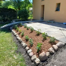 Ułożona kostka brukowa przed budynkiem z nowo zasadzonymi krzewami w korze, obramowanymi kamieniami.