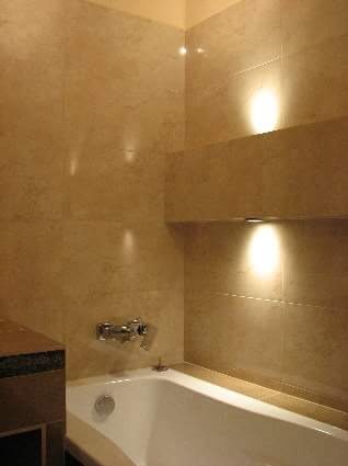 Wykończona łazienka z beżowymi płytkami imitującymi kamień, biała wanna z chromowaną baterią, oświetlenie punktowe w półce ściennej.