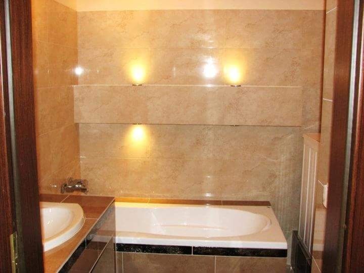 Wykończona łazienka z beżowymi płytkami imitującymi marmur, podświetlanymi niszami, białą wanną z czarnym obramowaniem i umywalką z brązowym blatem.