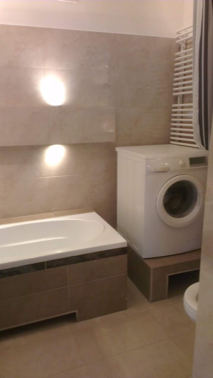 Jasna łazienka z wanną obudowaną brązowymi płytkami, białą pralką na podeście i grzejnikiem drabinkowym.