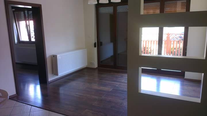 Wnętrze po remoncie: ciemna drewniana podłoga, białe ściany z grzejnikami, okna z roletami, widok na balkon przez otwory w ścianie działowej.