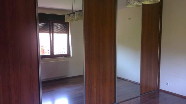 Wnętrze pokoju z drewnianą podłogą i przesuwnymi drzwiami szafy z lustrami, widok na okno z roletami i grzejnik.