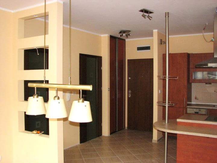 Wnętrze mieszkania po remoncie: widok na kuchnię z barkiem, oświetlenie sufitowe z trzema kloszami, brązowe drzwi i płytki podłogowe w tonacji beżowej.