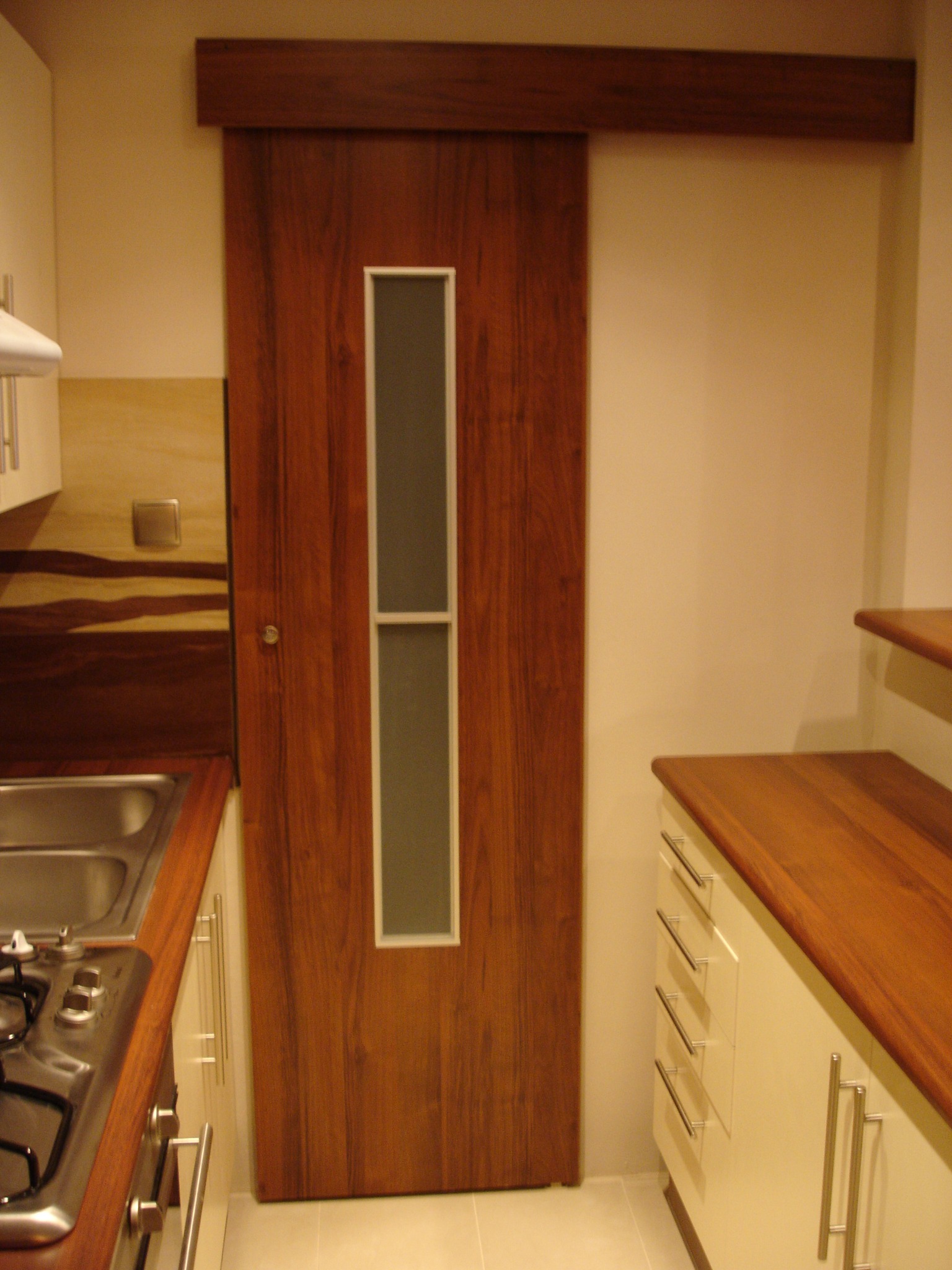 Brązowe, przesuwne drzwi z długim, pionowym przeszkleniem w jasnej kuchni z kremowymi szafkami i drewnianym blatem.