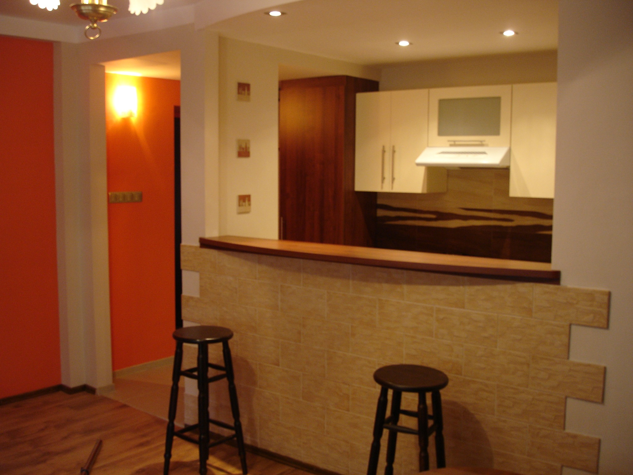 Wykończona kuchnia z niską ścianką działową wyłożoną beżowymi płytkami imitującymi cegłę, z dwoma drewnianymi stołkami barowymi, widok na pomarańczową ścianę w drugim pomieszczeniu.