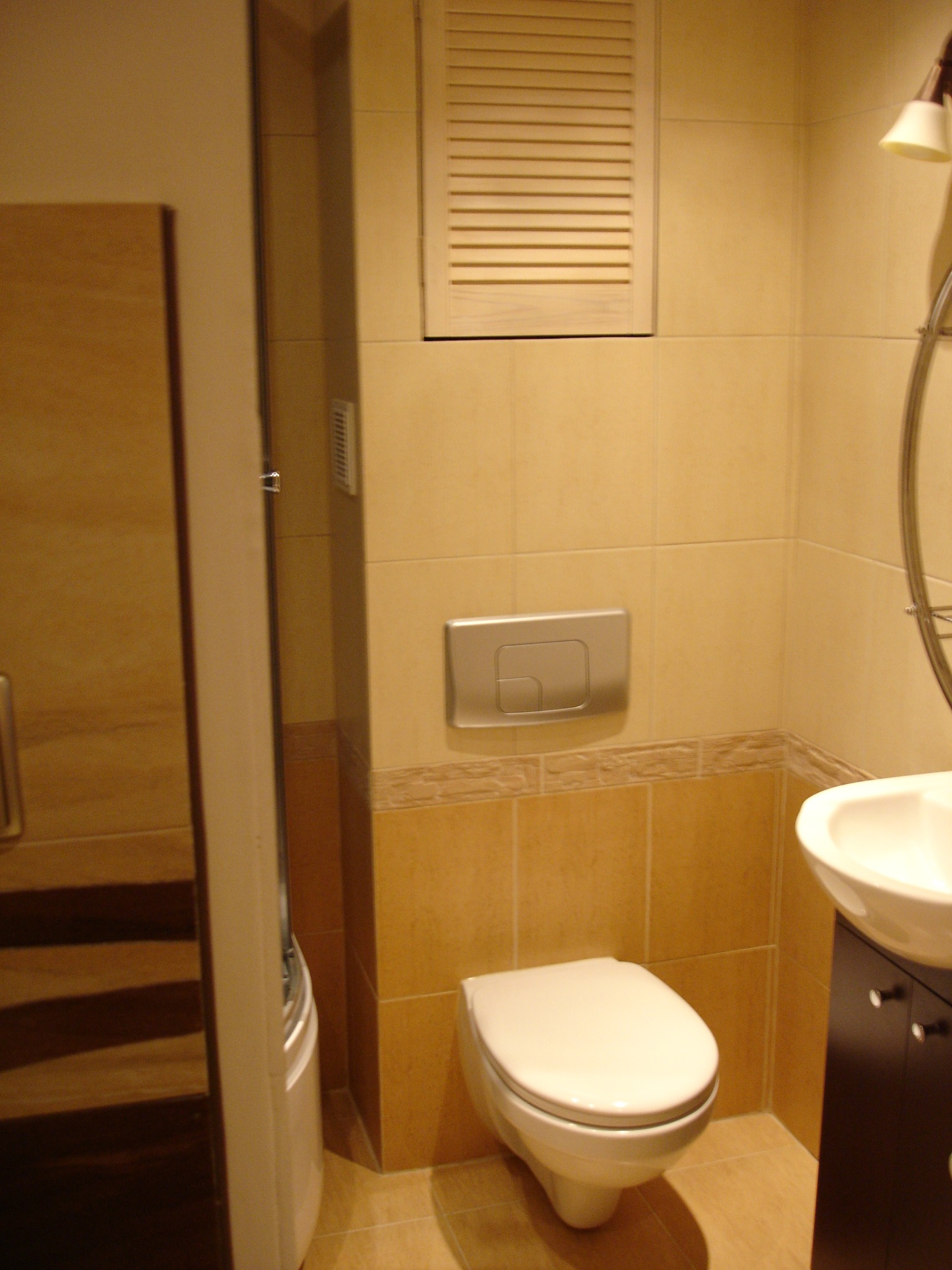 Jasna łazienka z beżowymi płytkami, wiszącą toaletą z chromowanym przyciskiem spłukiwania, umywalką z szafką i drewnianą szafką z lustrem, oświetlona lampą.