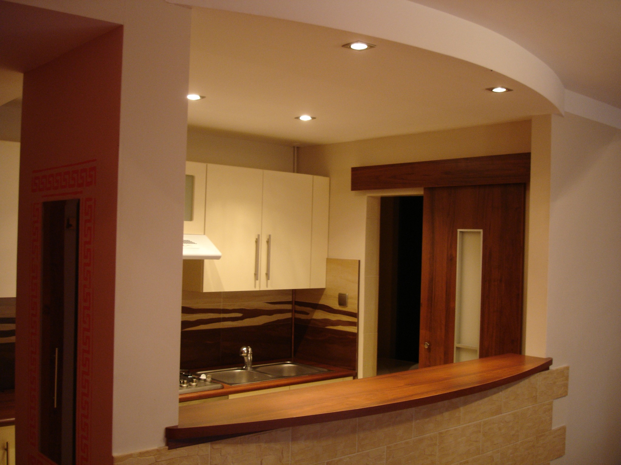 Wykończona kuchnia z drewnianym blatem i przesuwnymi drzwiami, widok na zlew i szafki, oświetlenie punktowe w suficie podwieszanym.