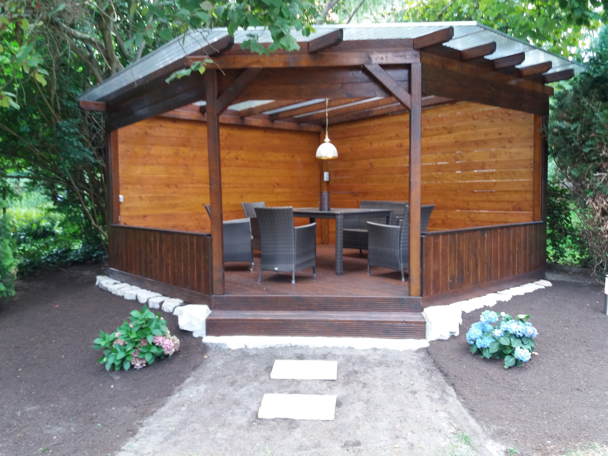 Drewniana altana ogrodowa z meblami rattanowymi, oświetlona wiszącą lampą, otoczona świeżo posadzoną zielenią i kamienną ścieżką.