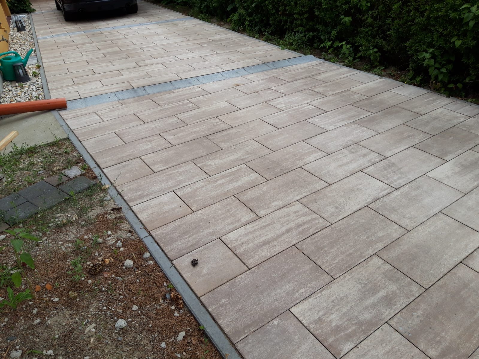 Ułożona kostka brukowa w odcieniach beżu i szarości, tworząca podjazd z widocznym odpływem liniowym. Z boku widoczne krzewy i trawnik z elementami ściółki leśnej.