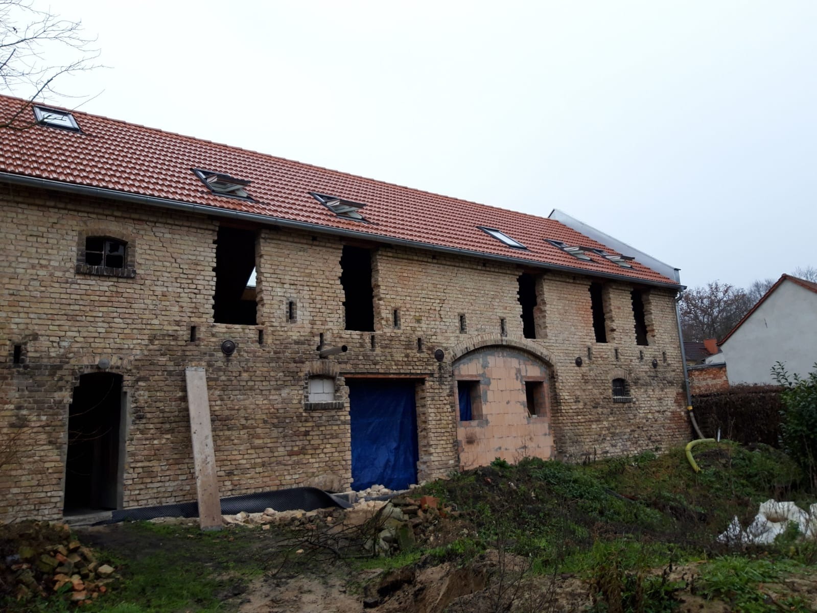 Budynek z żółtej cegły w trakcie remontu, z widocznymi otworami okiennymi i drzwiowymi, częściowo zabezpieczonymi niebieską plandeką, z czerwoną dachówką i oknami dachowymi, otoczony ziemią...