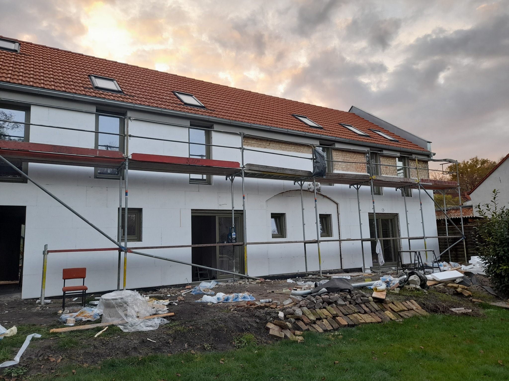 Dom w trakcie ocieplania elewacji, widoczne rusztowania, styropianowe płyty izolacyjne, cegły i krzesło na trawniku, pochmurne niebo w tle.