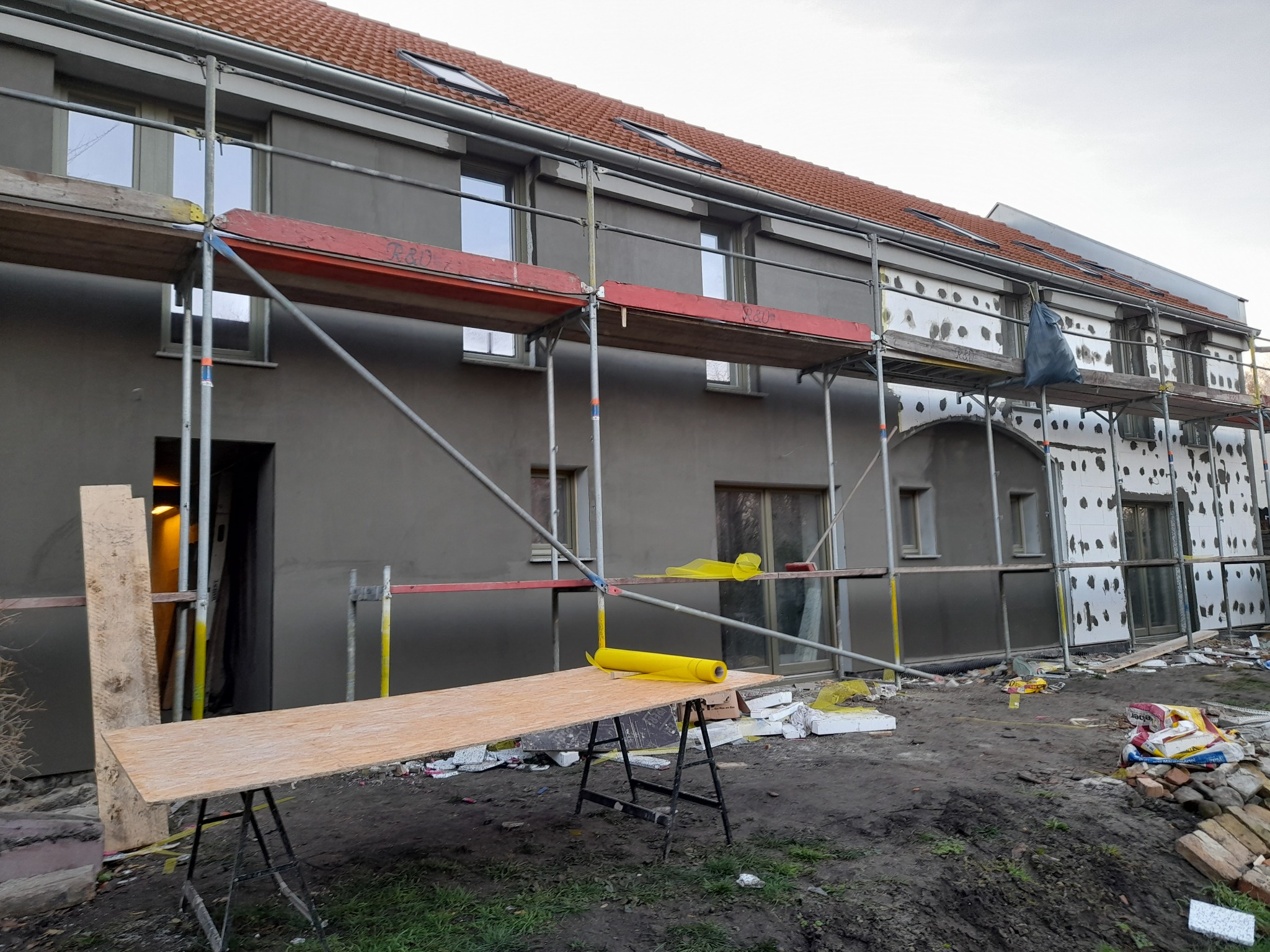 Budynek dwukondygnacyjny w trakcie prac elewacyjnych: częściowo ocieplony styropianem, częściowo pokryty szarym tynkiem; rusztowania, deska na kozłach z żółtą folią, bałagan na ziemi.