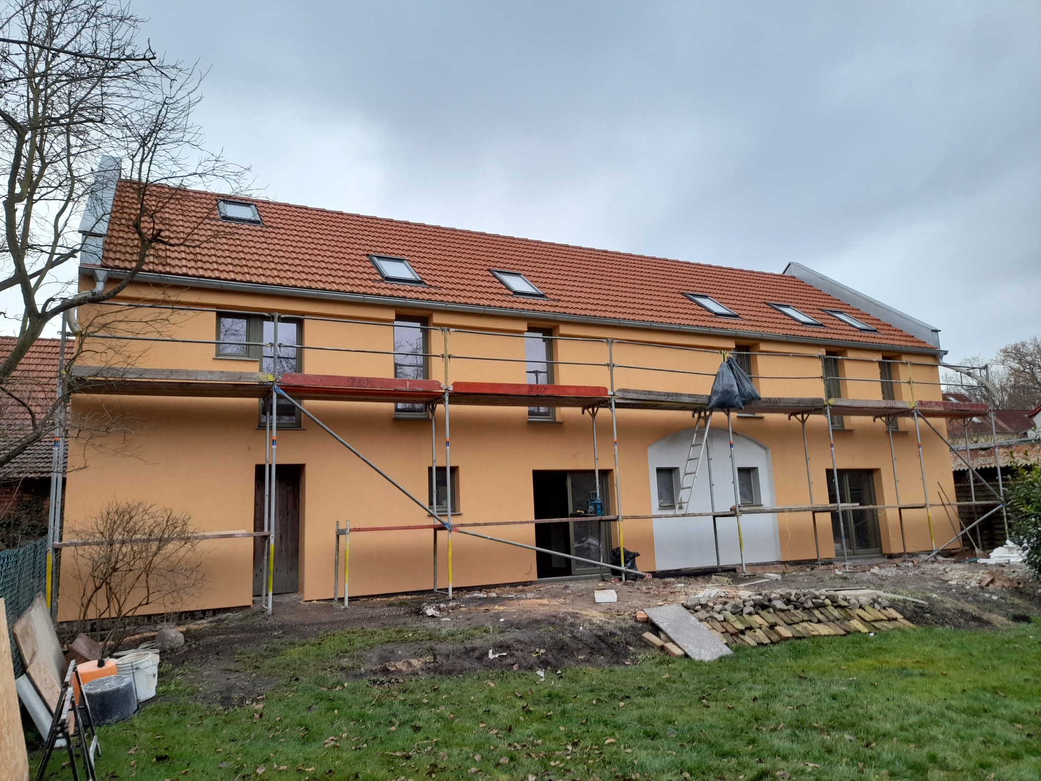 Dwukondygnacyjny budynek z pomarańczową elewacją w trakcie prac wykończeniowych, widoczne rusztowania, dach z dachówką i oknami połaciowymi, teren wokół częściowo zagospodarowany, częściowo z gruzem.