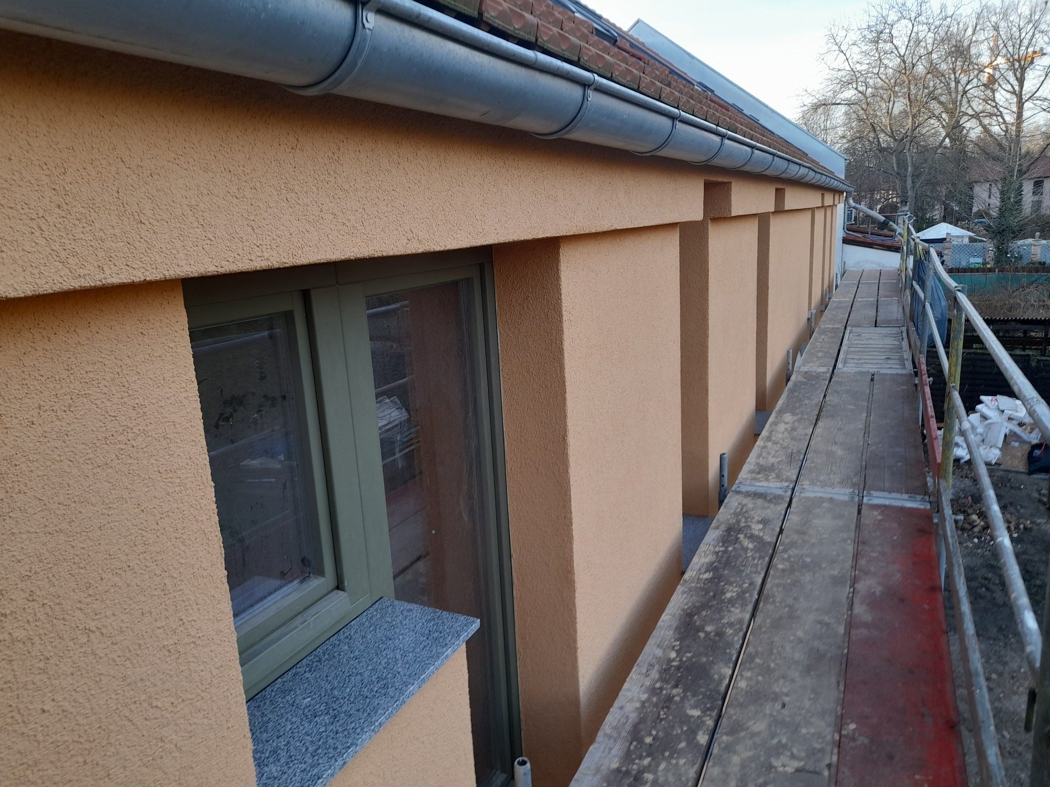 Fragment elewacji budynku z oknem w zielonej ramie i szarym parapetem, widoczny z rusztowania, z fakturą baranka w odcieniach beżu i pomarańczy.