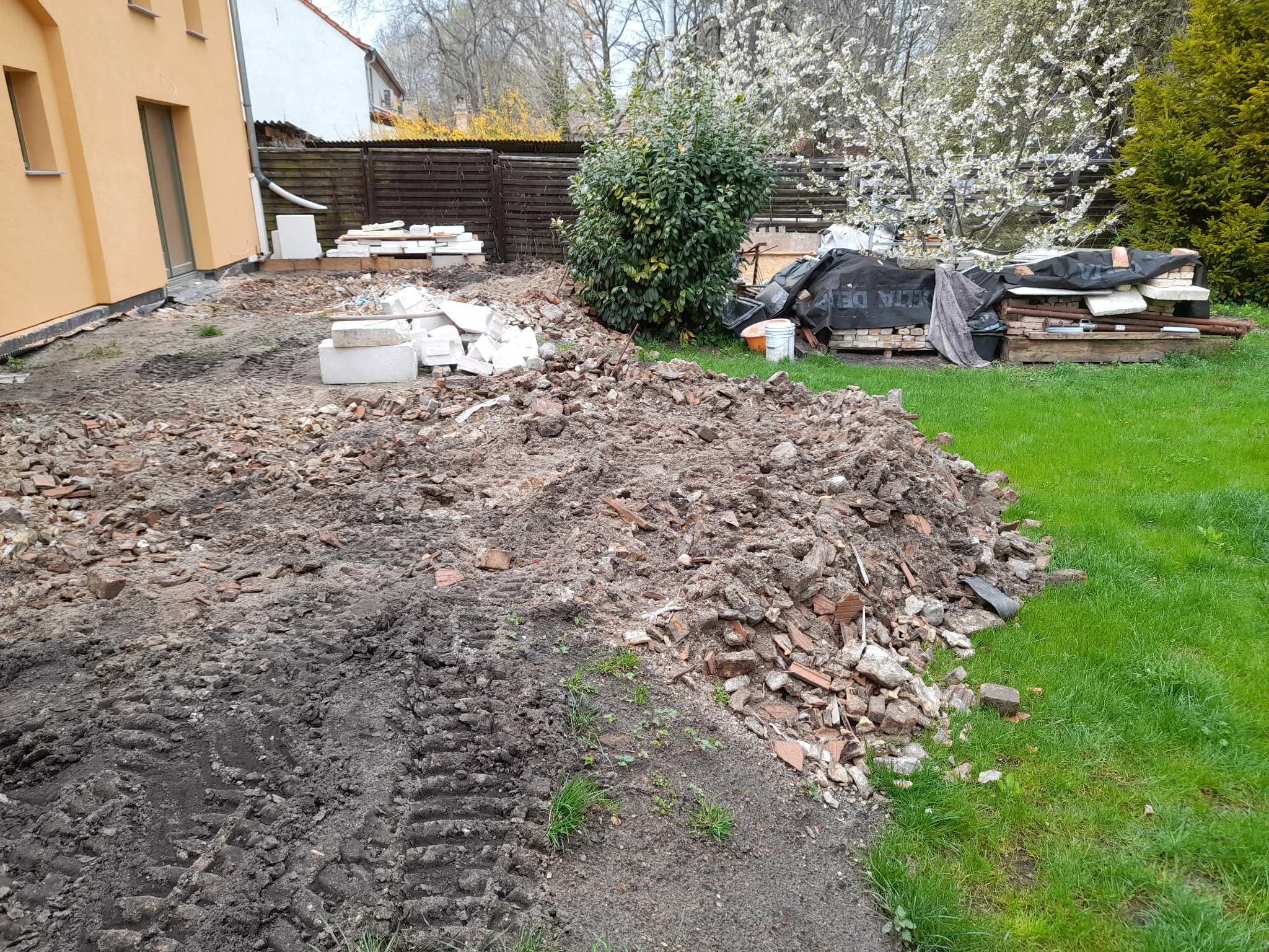 Teren po pracach budowlanych: sterta gruzu ceglanego i ziemi z wyraźnymi śladami opon, obok trawnik, w tle budynek z ocieploną elewacją, składowane materiały budowlane pod plandeką i kwitnące drzewo.